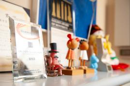 棚に賞状や受賞トロフィーが並び、手前にキャラクターのフィギュアが数体飾られているオフィスの一角