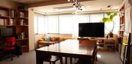 大きな木製のテーブルと木製チェア、ベージュのソファ、大型テレビが配置されたガレージフィルムの打ち合わせスペース。両脇の本棚には書籍やフィギュア、観葉植物が並ぶ