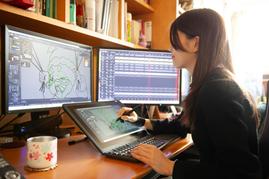 黒髪の女性がペンタブレットで作画しながら、2台のモニターに表示されたアニメーション制作ソフトを見ている横顔